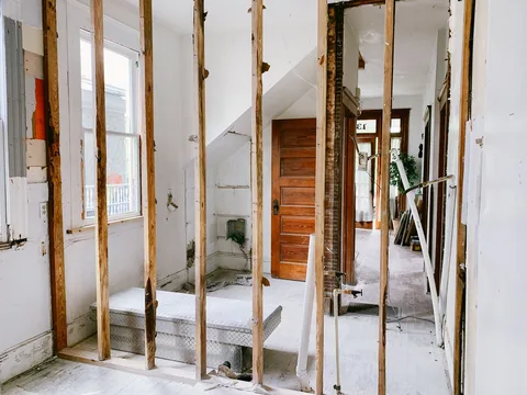 A room stripped to the wood studs mid-renovation, with plumbing lines exposed and a hallway beyond